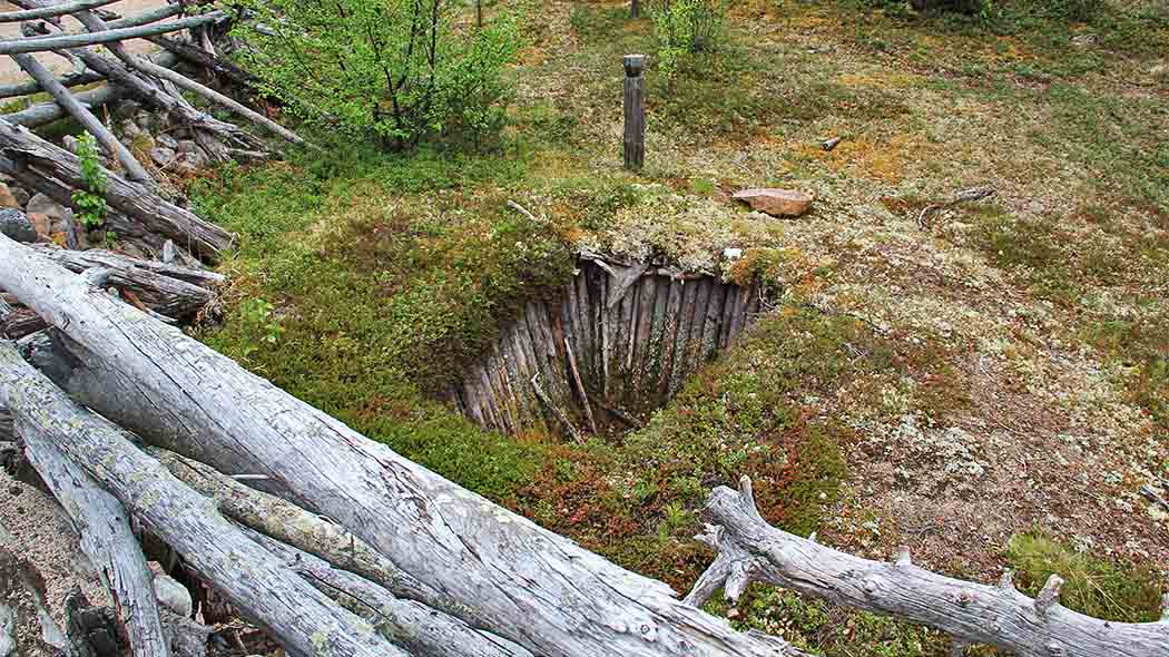 Keskellä kuvaa maassa kuoppa, jonka reunoilla puuseipäitä. Kuopan ympärillä jäkäläinen maa.