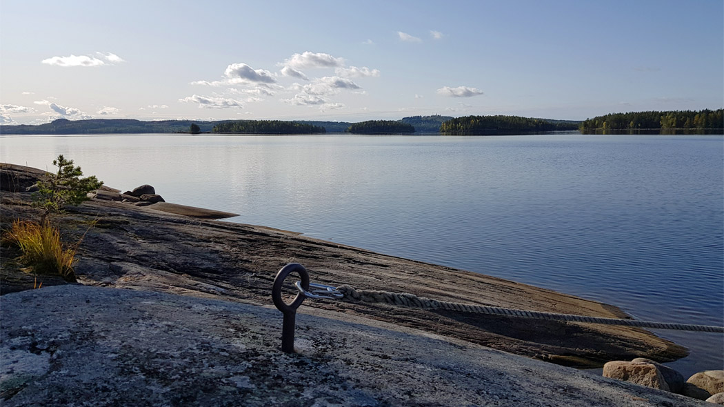 Lähes tyyni ja poutainen järvi - saaristomaisema alkuillan valossa. Etualalla on loiva, matala kallio, jossa on kiinnityspiste veneille.