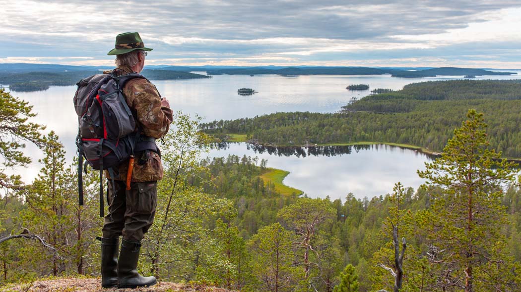 Retkeilijä seisoo reppu selässään korkealla mäellä näköalapaikalla. Mäeltä avautuu maisema jossa on vehreää kesäistä metsää ja järviä. Taivas on pilvinen.