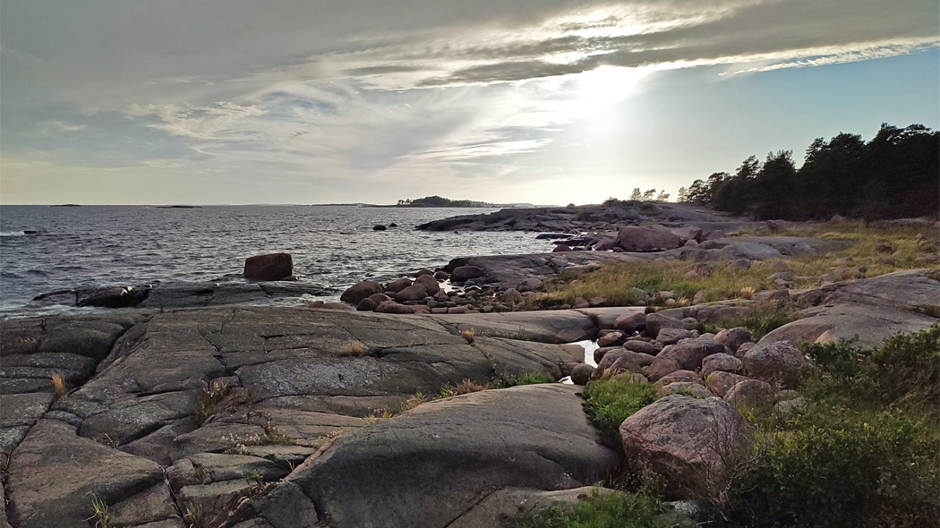 Saaristomaisema. Etualalla kalliota. Taaempana rantametsää, kallioista merenrantaa ja horisontti. Aurinko on pilven takana.