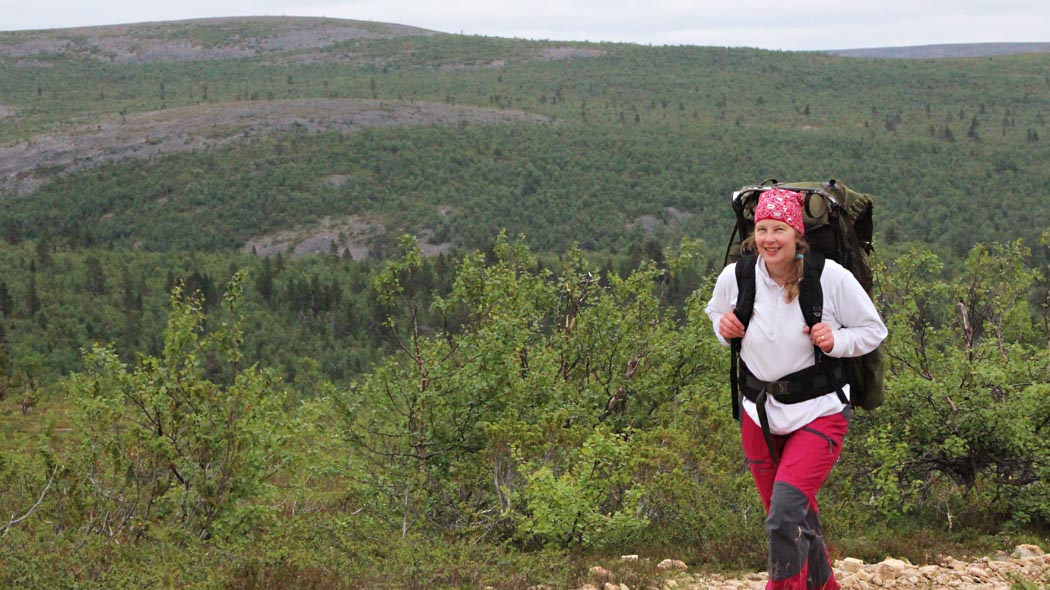 Etualalla nainen kävelee rinkka selässään pienten koivujen reunustamaa polkua. Taustalla on metsäisinä kohoavat tunturin rinteet sekä puuton tunturin huippu.