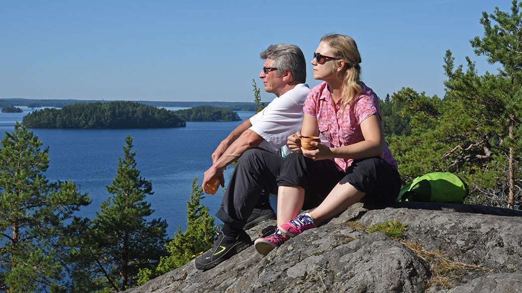 Kaksi retkeilijää istuu kalliolla. Taustalla järvimaisema.