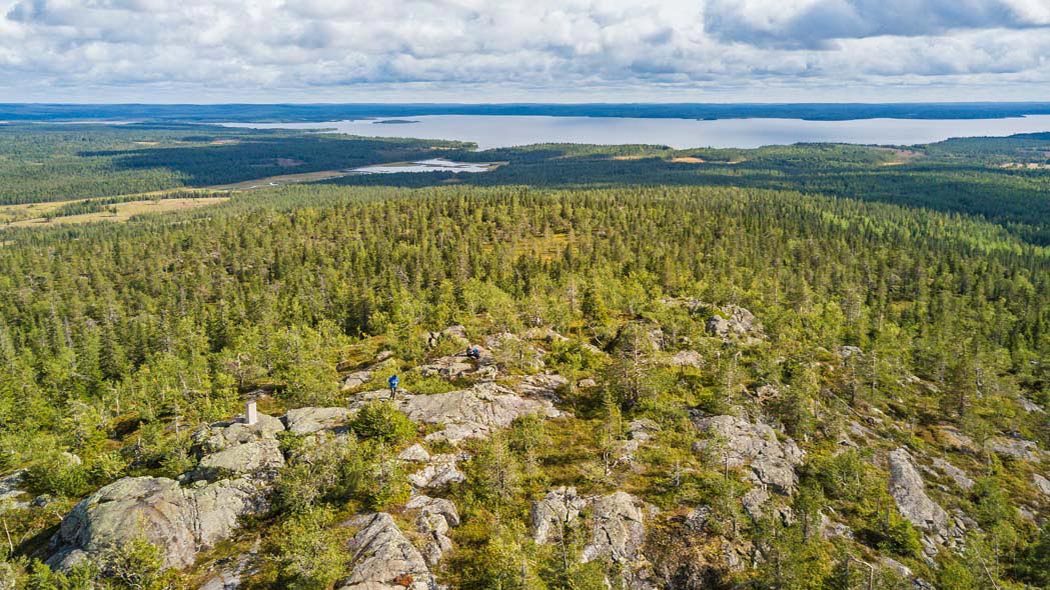 Ilmakuva Pyhityksen huipulta kohti Kostonjärveä. Edessä näkyy Pyhitystunturi, minkä huipulla seisoo sinitakkinen henkilö. Taustalla välkkyy Kostonjärvi ja vihreää metsää.