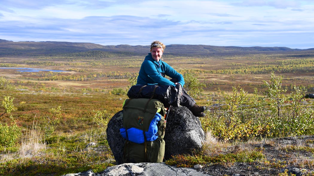 Vaeltaja lepää isolla kivellä istuen. Taustalla siintää avara tunturimaisema.