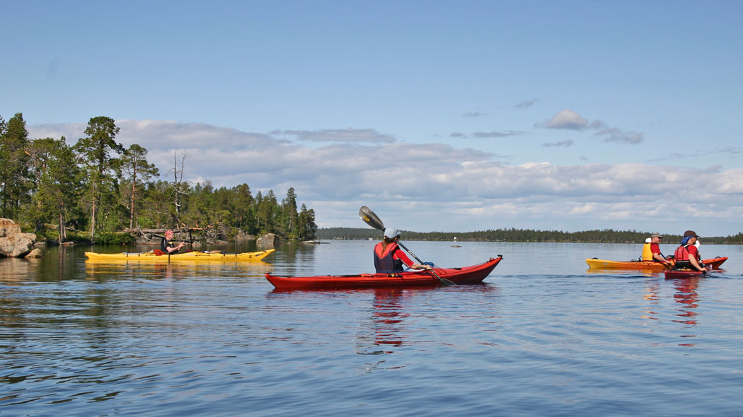 Retkeilijöitä melomassa järvellä puolipilvisenä kesäpäivänä. Järven rannassa kasvaa havumetsää.