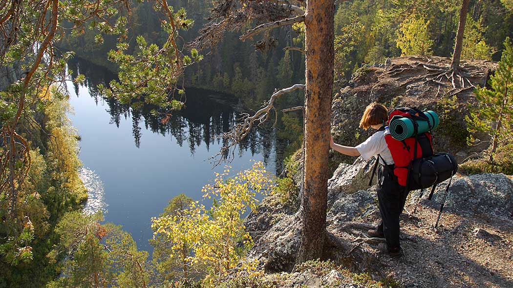 Retkeilijä seisoo rinkka selässä korkealla jyrkänteellä. Alhaalla virtaa joki kesäisen metsän keskellä.