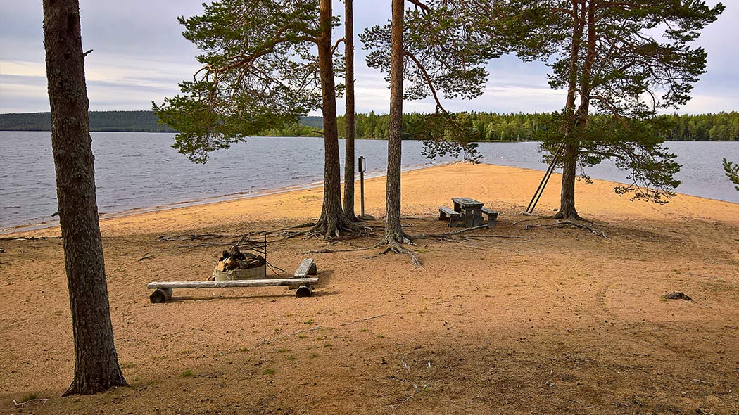 Hiekkaniemi, jossa on tulentekopaikka ja kasvaa muutama puu. Niemeä ympäröi järvi ja taustalla näkyy metsikköä. Orhinselän taukopaikan upea hiekkaniemi Miekojärvellä. Kuva: Anna Pakkanen