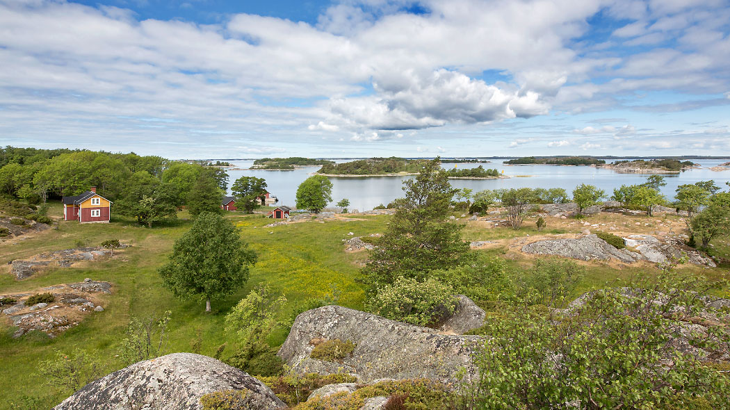 Kesäinen saaristomaisema. rantakalliolla on muutama puurakennus sekä kasvillisuutta. Kuva: Jari Kostet.