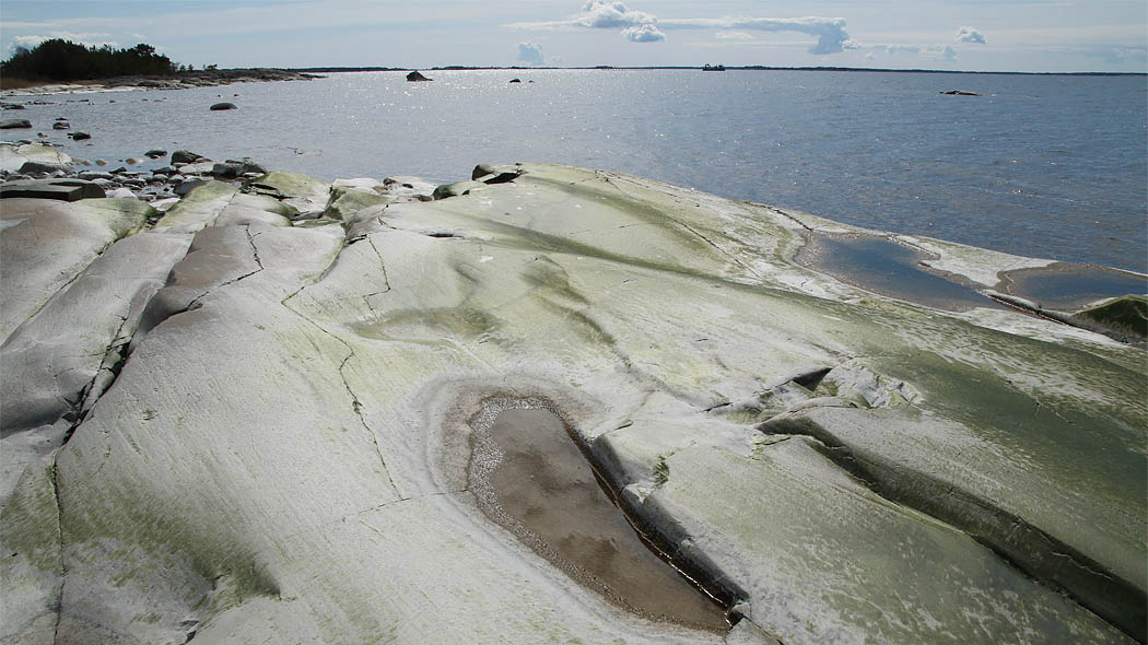 Etualalla sileä rantakallio, jonka takana avoin vesialue ja taivas pilvineen.