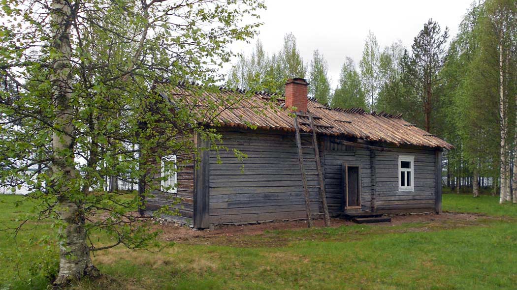 Pihapiirin keskellä harmaa rakennus. Taaempana näkyy puita, joiden lävitse näkyy sininen järvi. Vasemmalla vihreälehtinen koivu.