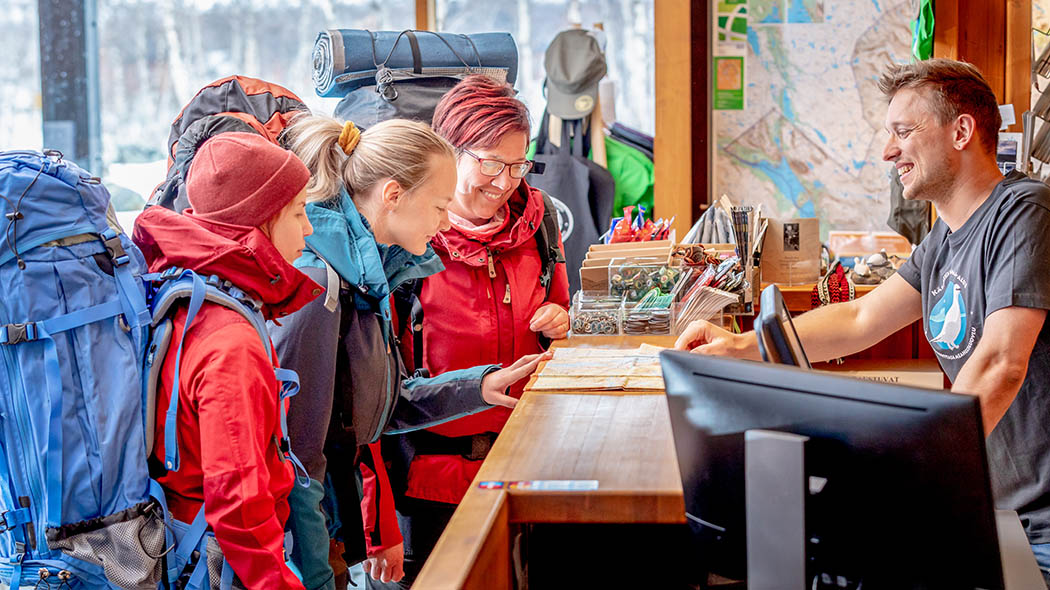 Kolme retkeilijää rinkkojen kanssa on saapunut luontokeskuksen palvelutiskille.