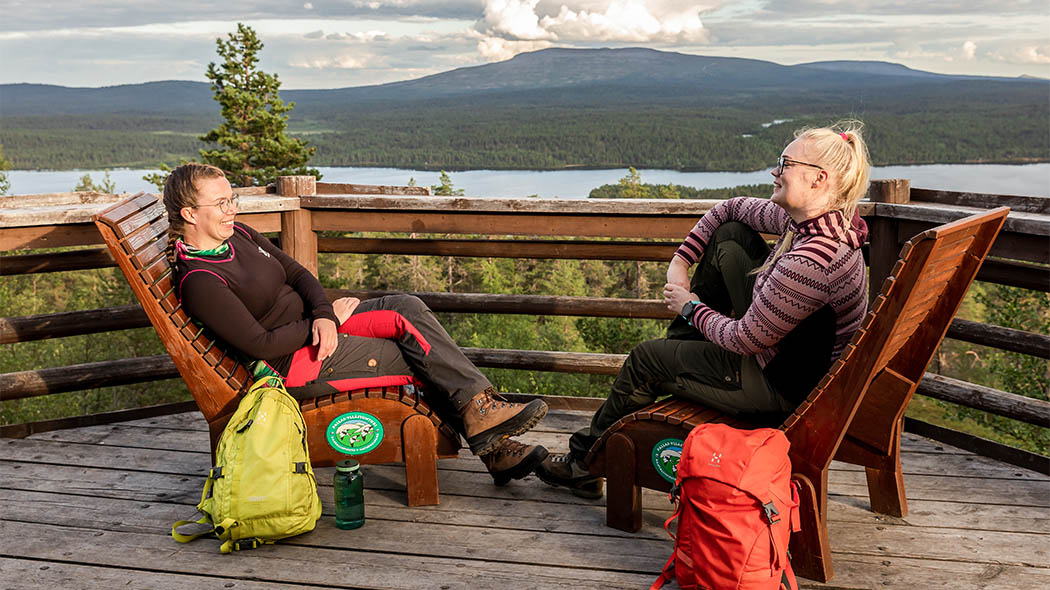 Kaksi hymyilevää naista istuu vastakkain puisilla lepotuoleilla katseluterassin kaiteen vieressä. Taustalla näkyy kesäinen maisema tunturiin. Retkeläisten reput ovat maassa tuolien vieressä.