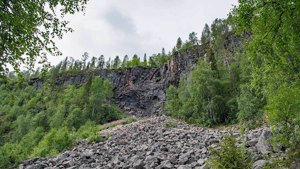 Suuri kallioseinämä, jonka alapuolella paljon kivilohkareita. Kallioseinämää reunustaa vihreät lehtipuut.