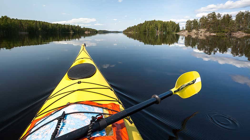 Kuvan etualalla on mela ja kanootin keula, jonka kannella on kiinnitettynä kartta. Järven rannoilla on metsiä ja kallioita.