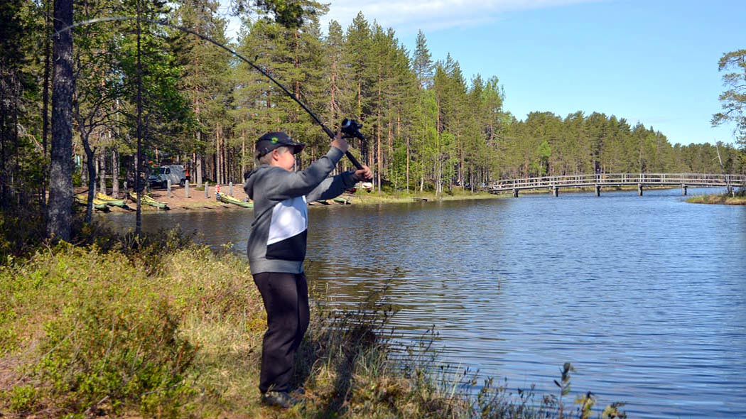 Lapsi heittää viehettä virvelillä järvenrannassa. Järven yli menee kapeasta kohdasta puinen kaarisilta. Vastarannalla on veneranta ja auto.