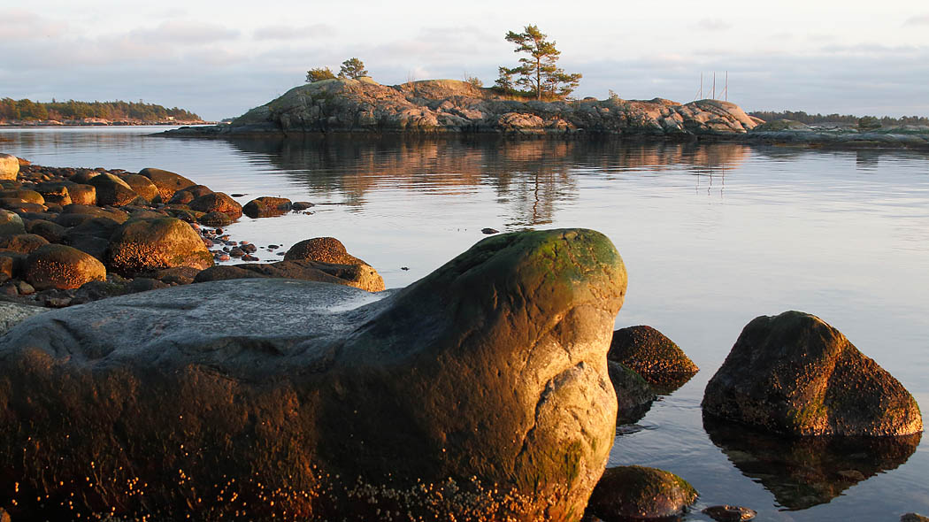 Syksyinen merenranta auringon laskiessa. Etualalla kiviä, taaempana kivinen pieni saari.